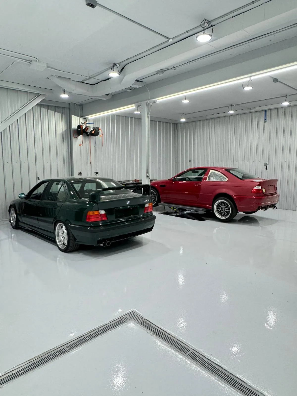 British Racing Green E36 alongside Imola Red E46 on the clean shop floor. Two builds in progress.